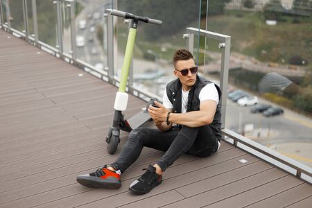 Handsome young man model in sunglasses in a trendy denim vest in jeans in sneakers with a phone sit...