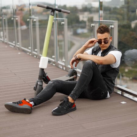 Cool american hipster man in fashionable denim black clothes in sunglasses sit on the wooden floor...
