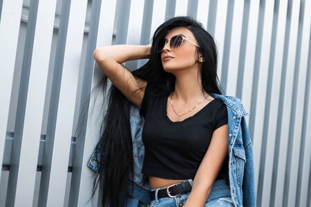 Young hipster woman with luxurious long black hair in a stylish top in a fashionable denim jacket in sunglasses relaxes in the city near a metal wall. American trendy girl fashion model. Street style.の写真素材