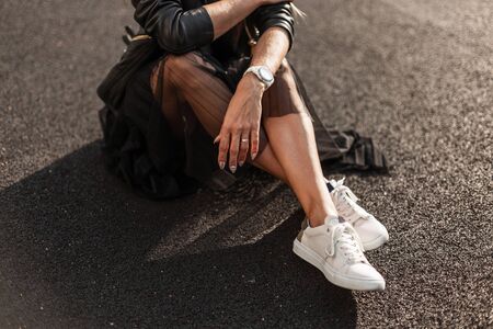 Young woman in stylish white sneakers in a vintage black skirt in a trendy leather black skirt sits on the asphalt on a sunny day. Fashionable autumn collection of youth women's clothing. Close-up.の写真素材