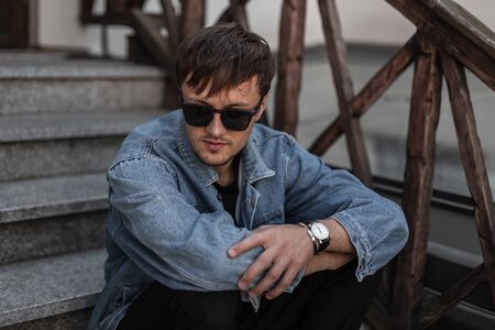 Stylish cool young man hipster in blue denim clothes in trendy sunglasses sits on a vintage...
