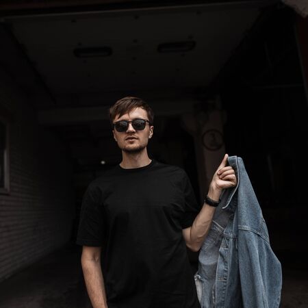 Pretty fashionable young man hipster with a beard with a stylish hairstyle in a trendy black T-shirt in sunglasses with a denim jacket posing in a dark building. Attractive brutal guy fashion model.の写真素材
