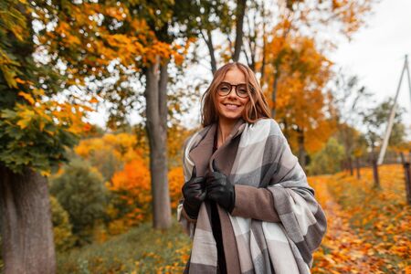 Joyful young hipster woman in a fashionable knitted scarf in stylish glasses in a trendy coat posing in a park outside the city. Cheerful girl model with a positive smile walks in the forest. Autumn.の写真素材