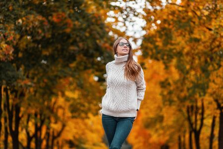 Joyful young woman model in a white knitted sweater in stylish glasses stands in the park and smiles...