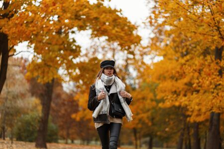 Pretty trendy young woman in a fashion hat with a bag in a vintage jacket in leather leggings with a scarf travels through the autumn park on the background of trees with golden foliage. Girl outdoorsの写真素材