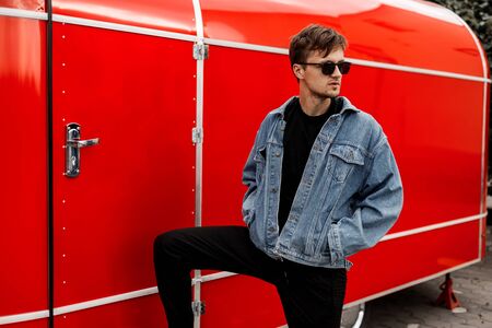 Fashionable young hipster man in a fashionable blue denim jacket in black pants in stylish sunglasses with a beard near a red metal van on the street. American guy resting on a summer day in the city.の写真素材