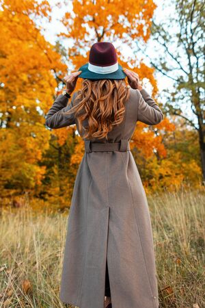 Back view of a fashionable young woman in a stylish long coat in an elegant hat with a trendy hairstyle in an autumn park among trees with orange foliage. Modern girl walks through the woods.の写真素材
