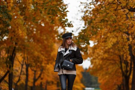Modern European young woman in stylish warm clothes in a vintage hat with a black leather bag travels in a autumn park. Pretty trendy girl fashion model in the forest on a background of gold leaves.の写真素材
