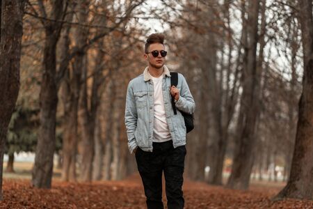 Trendy young hipster man with a stylish hairstyle in a vintage blue denim jacket in pants in sunglasses with a backpack travels on the autumn park on a October day.の写真素材