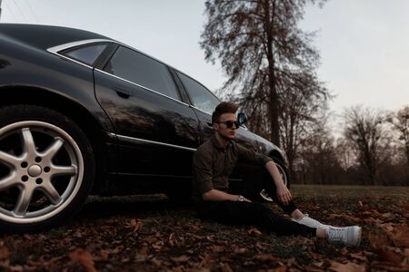 Young successful handsome man hipster in stylish clothes in trendy sunglasses in fashionable sneakers sits on dry golden foliage near a modern car in the park.の写真素材