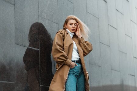 Cute sexy young blonde woman in a white vintage sweater in a stylish beige trench coat in jeans posing in the city near a black modern building.の写真素材