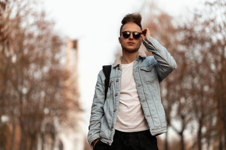 Stylish young man with a beard with a fashionable hairstyle in trendy sunglasses in a blue denim...