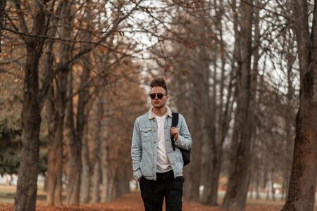 European trendy handsome young man in stylish jeans clothes in vintage sunglasses with a fashionable hairstyle with backpack walks on the autumn forest.の写真素材