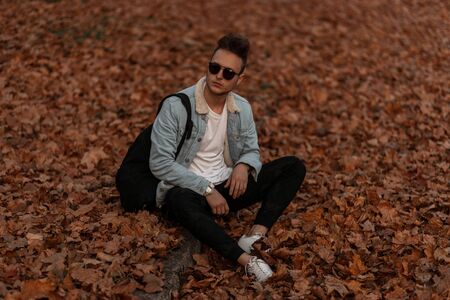 Stylish young man with a hairstyle in sunglasses in military pants in white sneakers in a denim jacket with a backpack sits on golden foliage in the park.の写真素材