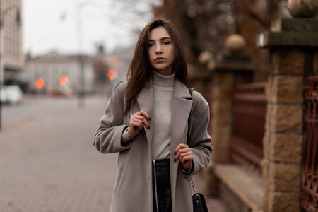 Model of a young woman in elegant outerwear with a black leather bag posing outdoors in the city. Attractive trendy girl fashion model enjoys walking on the street. Fashionable women's autumn clothes.の写真素材
