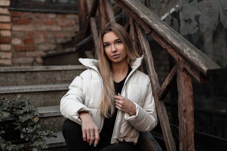 Glamorous cute young blond woman in a trendy white jacket in black stylish jeans sits on a stone staircase in the city. Beautiful  modern pretty girl fashion model resting outdoors. Autumn style.の写真素材