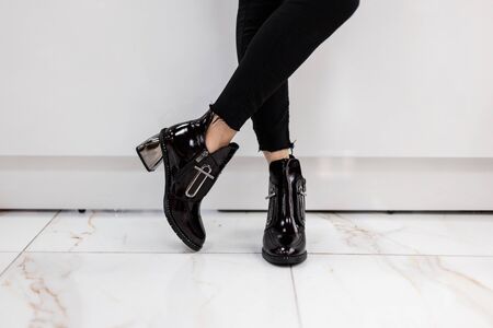 Fashionable young woman in stylish black jeans in trendy patent leather shoes with silver heel stands near a white wall in a shopping center. Girl chooses autumn boots. Shopping. Close-up.の写真素材