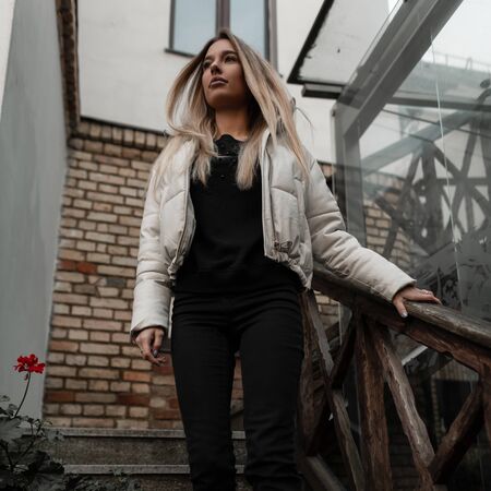 Glamorous cute young blonde woman in a fashionable white jacket in black stylish jeans descends down a vintage staircase. Beautiful modern pretty girl fashion model outdoors. Spring-autumn style.の写真素材