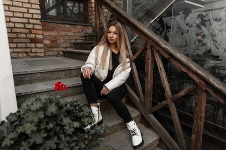 Fashionable cute young woman model in a white stylish jacket in black jeans in leather white trendy boots sits on an old stone staircase in the city. Attractive modern girl outdoors in a autumn day.の写真素材