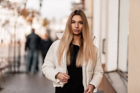 Glamorous young woman blonde with sexy lips in a black stylish sweater in a fashionable white winter jacket walks outdoors near a vintage building. Cute girl model stands in street. Youth fashion.の写真素材
