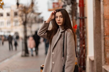 Young beautiful woman fashion model with long brown hair in a fashionable gray autumn coat with a leather black handbag posing near a vintage building in the city.Attractive girl resting on the streetの写真素材