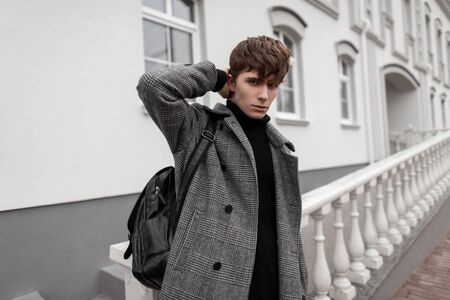 Sexy guy in stylish autumn-winter outerwear straightens hair. European young attractive man model in a gray plaid coat with a fashionable leather backpack poses outdoors near a vintage white building.の写真素材