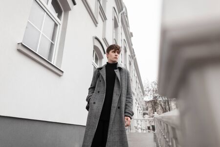Handsome young man in black clothes in an elegant gray plaid coat with a trendy hairstyle walks on...
