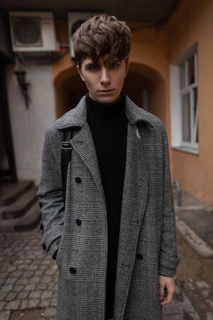 Young man model in an elegant gray plaid coat in a black knitted sweater with a trendy hairstyle posing in the city in the courtyard of a vintage building. Nice guy in stylish clothes outdoors. Style.の写真素材