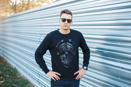 Attractive young hipster man in black trendy sunglasses in fashionable black shirt with pattern...