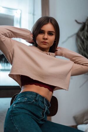 Fashion model of a young woman in a fashionable pink sweatshirt in stylish vintage jeans sits on a wooden chair in a cozy bedroom with a bed and a vintage mirror with bright lamps. Glamorous girl.の写真素材