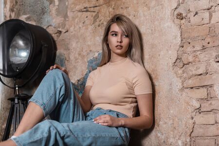 Slim European pretty young woman is resting near a brick wall in a vintage studio. Fashionable girl fashion model in youth casual wear relaxes near a black modern metal lamp. Trendy cute lady. Style.の写真素材