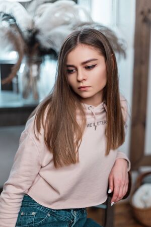 Portrait of a cute beautiful young woman in fashionable casual wear. Stylish girl fashion model with brown long hair in a pink sweatshirt is resting on a chair near a vintage mirror in a cozy bedroom.の写真素材