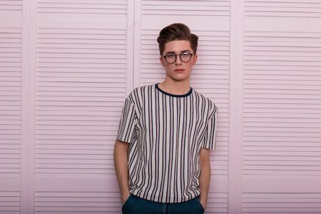 Modern sexy young man hipster in fashionable glasses with a hairstyle in a trendy striped t-shirt stand near a vintage wooden pink wall in the studio. Elegant handsome modern guy. English fashion.の写真素材