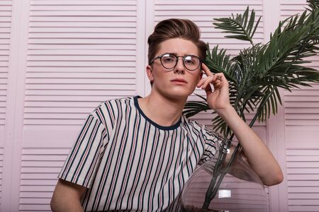 Portrait of a stylish young man hipster in a trendy striped T-shirt in jeans with glasses with a green exotic palm in a glass pot near pink wall in the studio. Sexy hipster guy posing in the room.の写真素材