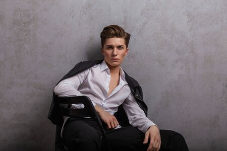 Young handsome man hipster in elegant white shirt in a fashionable leather black jacket is sitting...