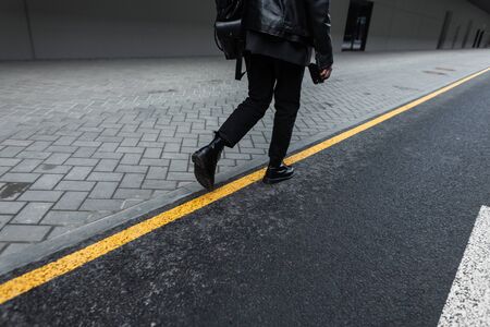 Back view on a fashionable young man in a black leather jacket in stylish jeans in leather boots with a trendy backpack in the city. Modern guy travels in fashion clothes on the street. Black style.の写真素材