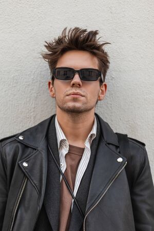 Fashionable portrait young man with serious face in a vintage dark sunglasses in a trendy leather black jacket with a stylish hairstyle near the wall outdoors. Handsome modern hipster guy in the city.の写真素材