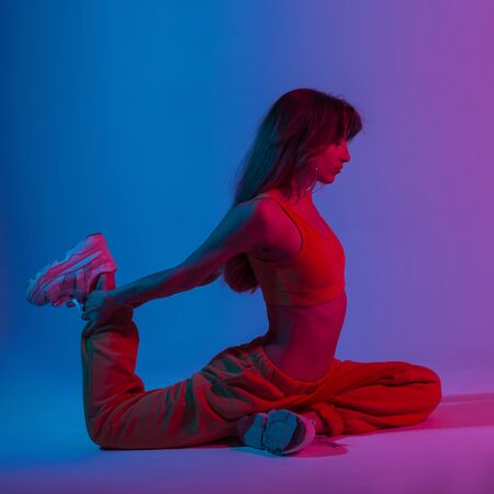 Young beautiful woman in a sexy top in pants doing yoga in a studio with bright neon blue-violet light. Sporty girl in trendy wear doing stretching with a multi-colored color in the style disco.の写真素材