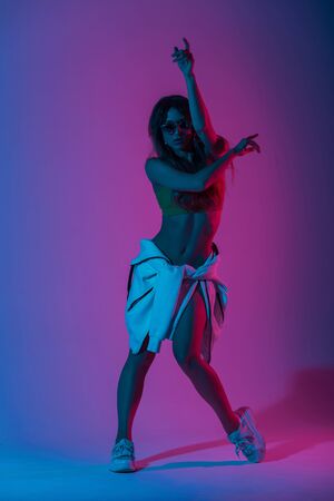 Pretty young woman dancer in a stylish top in a shoe in a hoodie dancing in the studio with a brightly neon blue light. Dance fashionable girl fashion model in sunglasses indoors with colored glitterの写真素材
