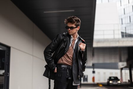 Handsome European young man in fashion youth oversized clothes with a trendy hairstyle is resting near a modern building in the city on a spring day. Modern guy hipster in stylish sunglasses outdoors.の写真素材