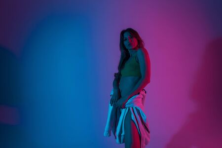 Modern sexy young woman in fashionable youth clothes posing indoors with neon violet-blue color in...