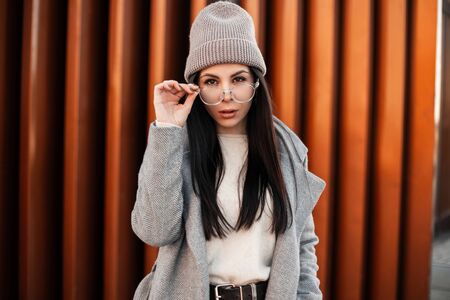 Urban trendy young hipster woman in stylish youth clothes straightens fashionable glasses and looks at the camera outdoors. Cute brunette girl in knitted hat in coat posing on the street in the city.の写真素材