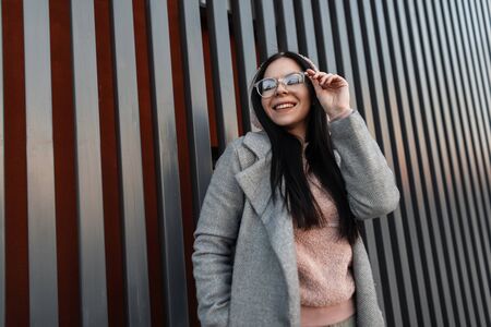 Happy young woman hipster straightens trendy glasses and positive smiles. Fresh portrait cute joyful girl with lovely smile in gray fashionable coat in vintage pink hoodie near metal wall in the city.の写真素材