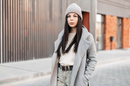 Pretty stylish young brunette woman in knitted hat in beige sweater in gray coat walks on street near vintage building on bright sunny day. Beautiful girl fashion model in casual wear. Street style.の写真素材