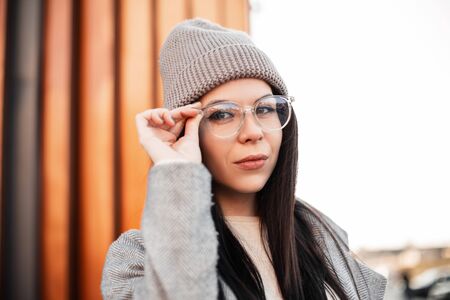 Stylish portrait pretty beautiful young woman brunette in fashionable knitted gray hat in vintage coat on the street. Attractive trendy girl hipster straightens fashion glasses. Casual spring look.の写真素材