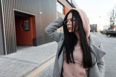 Attractive young woman model in pink trendy hoodie in fashionable glasses in stylish coat walks near vintage building on the street. Cute European hipster girl in the city. Casual clothes for women.の写真素材