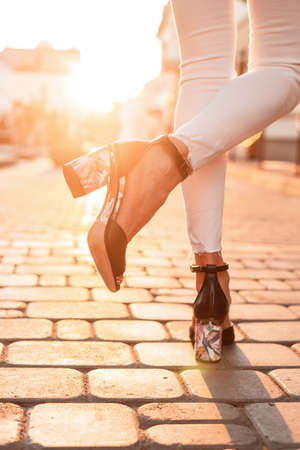 Close-up of female legs in leather black fashionable summer sandals in stylish jeans on a stone road in the city on a sunny dayの写真素材