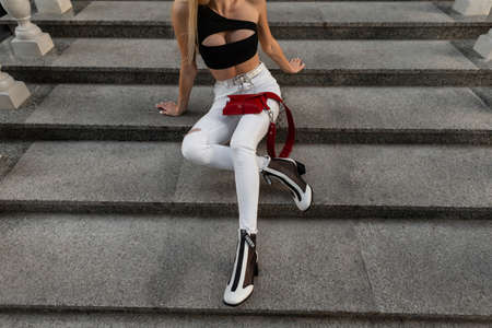 Young woman in fashionable denim clothes with a red bag and stylish leather shoes sits on the steps...