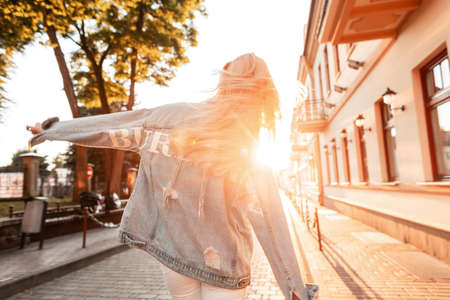 Pretty stylish blonde woman in vintage denim clothes dancing on the street at sunset. Back view.の写真素材