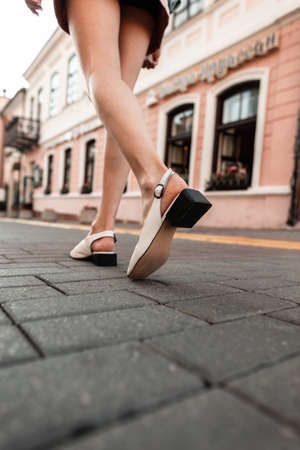Fashionable woman with pretty legs in stylish summer sandals walks on a stone road in the city. Back...